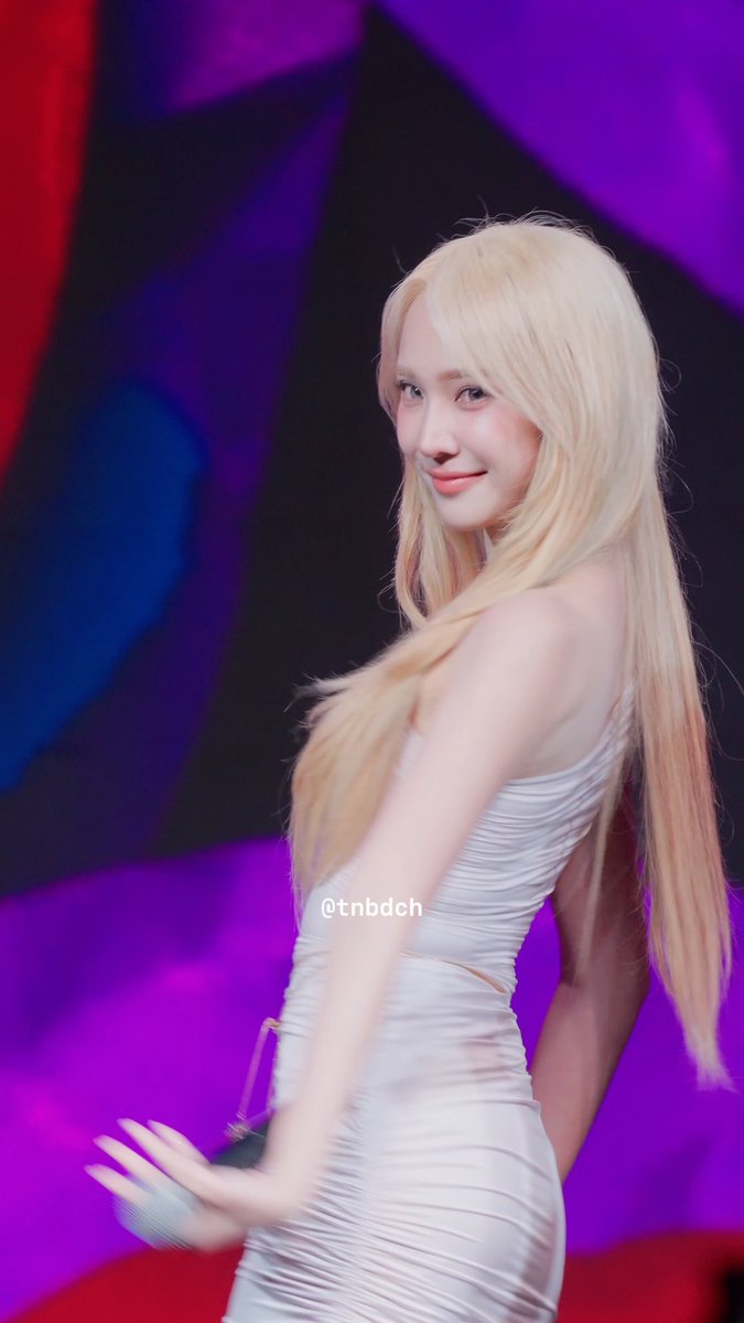 A blonde woman in a white dress turns and smiles at the camera against a vibrant purple and red stage backdrop, with the watermark '@tnbdch' visible.