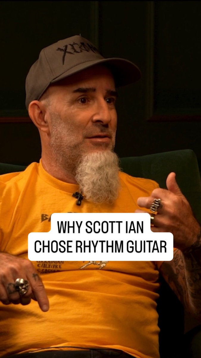 Scott Ian, wearing a gray cap and orange shirt with a distinctive white beard, sits for an interview with text overlay reading "WHY SCOTT IAN CHOSE RHYTHM GUITAR," referencing his discussion with Billy Corgan on The Magnificent Others about his preference for learning full songs rather than focusing on solos.