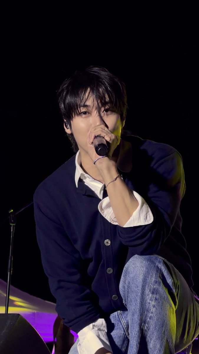 A young man with dark hair crouches on stage, gripping a microphone close to his face while singing, wearing a navy cardigan over a white collared shirt and blue jeans. The caption "261001 왕자님 얼굴 진짜 앙 그자체 #이승협" (translated: "261001 His Highness's face is truly adorable #LeeSeunghyup") fits the image perfectly, capturing the performer's endearing stage presence.