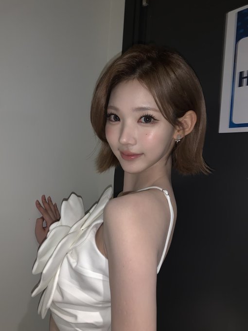 A close-up portrait of a young woman with short brown hair and a white ruffled top, posing indoors against a gray and dark wall.