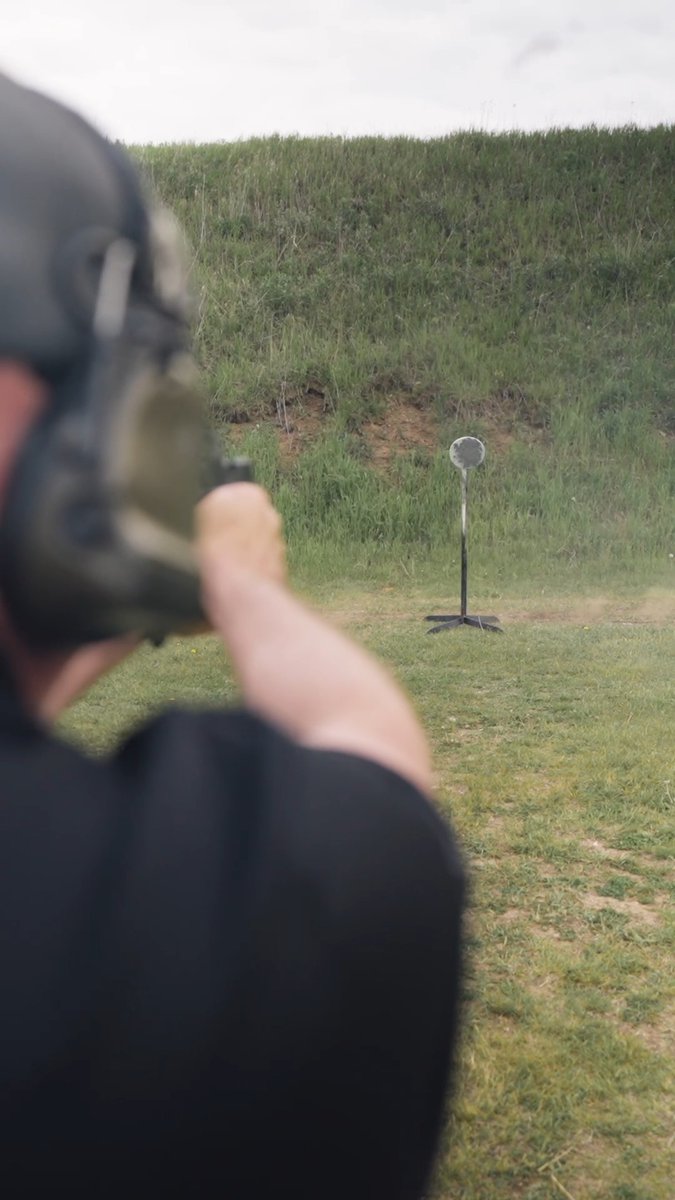 A person wearing ear protection fires a pistol at a steel target on a stand in an outdoor grassy field.