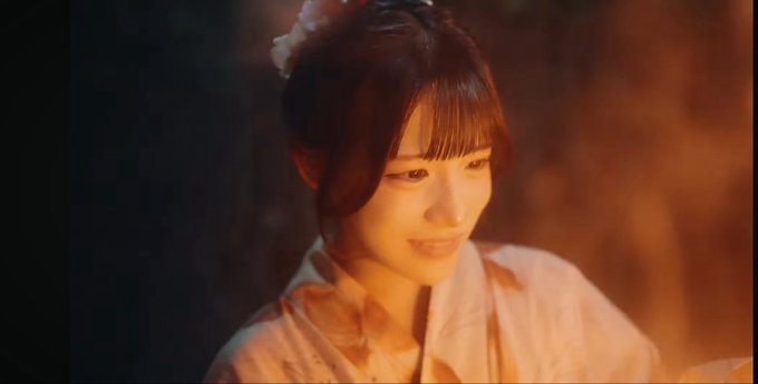 A young woman with dark hair styled in a traditional updo with bangs, wearing a light-colored garment, softly smiles in warm, dim lighting.