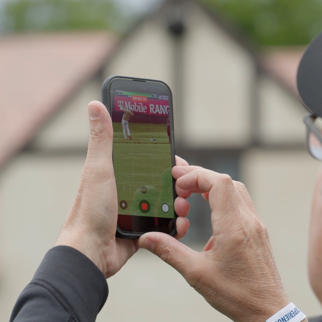 A person's hands hold a smartphone recording a T-Mobile Range golf event, with the camera interface showing the golfer mid-swing.