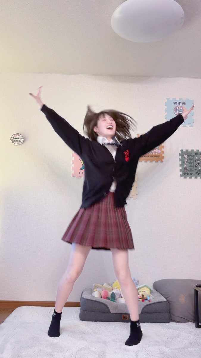 A photo of a young woman in a school uniform (black cardigan, white shirt, bow tie, and plaid skirt) jumping joyfully with her arms and legs spread, indoors in a room with a pet bed, toys, and wall decals including one that says "WAY BEYOND".