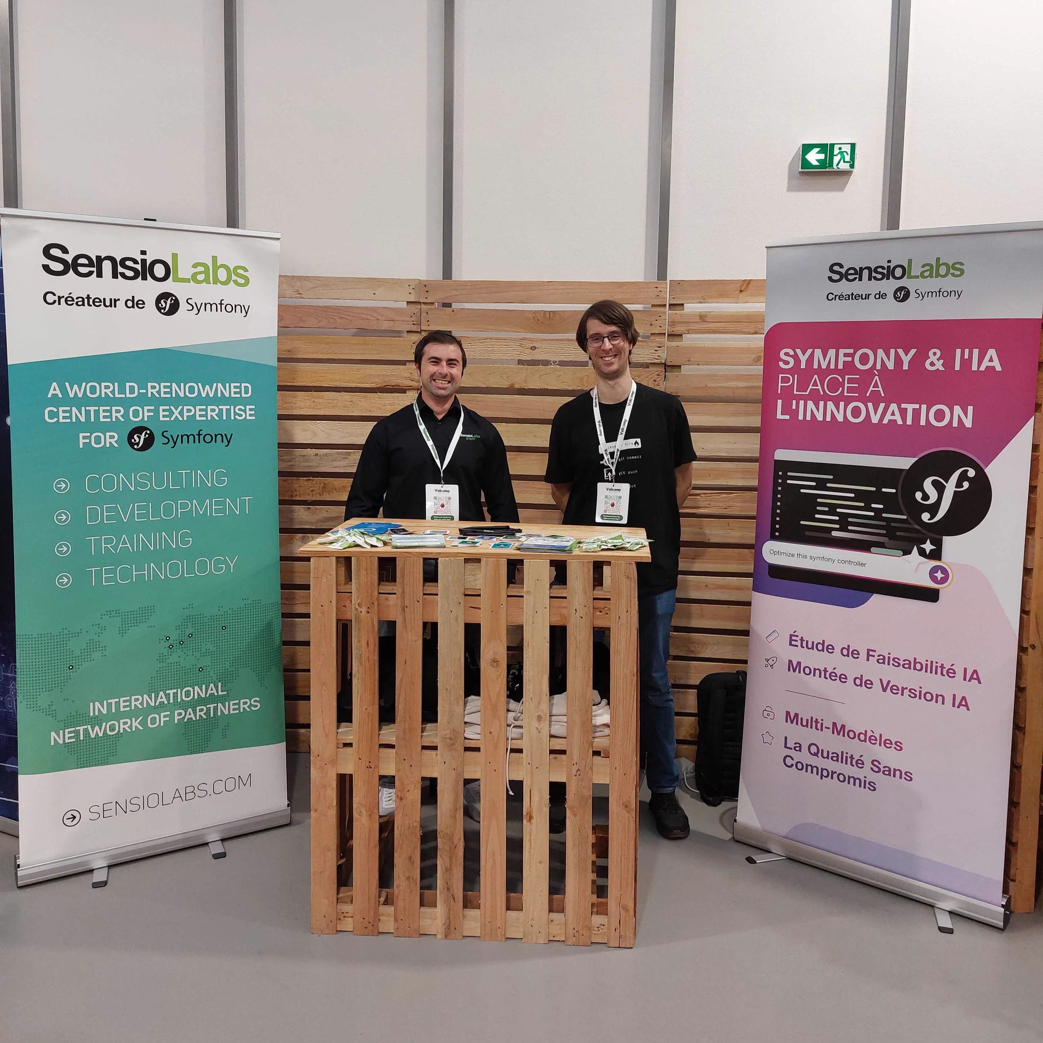 Le stand de SensioLabs avec 2 kakémonos (l'un sur Symfony et l'IA, l'autre sur l'expertise de SensioLabs), les deux membres de l'équipe qui tiennent le stand composé de palettes en bois