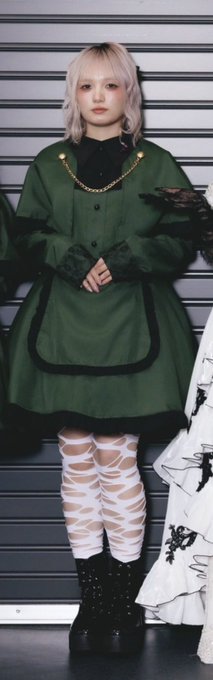 A performer poses in a green fur-trimmed dress over a black shirt, paired with white ripped tights and chunky black boots.