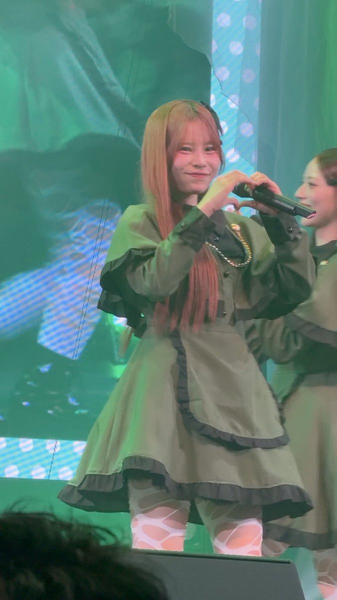 A young woman with long, straight hair performs on stage wearing an olive green dress, forming a heart shape with her hands while holding a microphone.