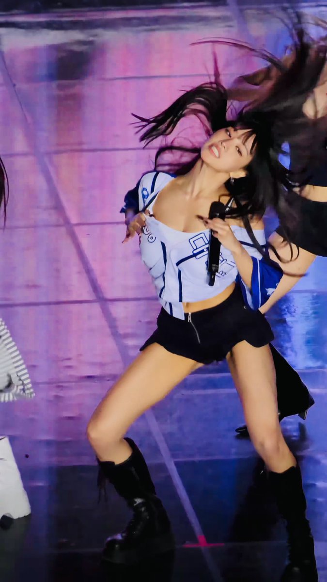 A K-pop female performer in a white and blue cropped jersey top and black skirt dances energetically on stage with a microphone in hand, her long hair flying.