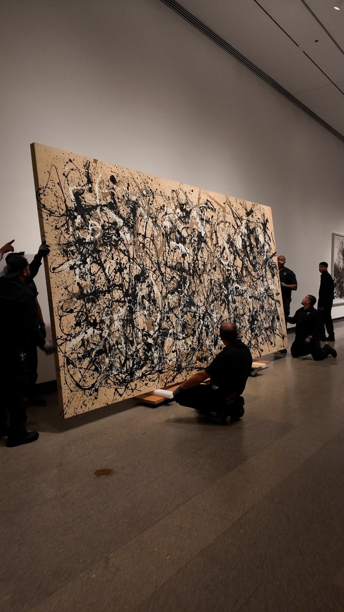 Art gallery scene where several workers in black uniforms are carefully installing or moving a large abstract expressionist painting featuring black and white drip patterns on a beige background.