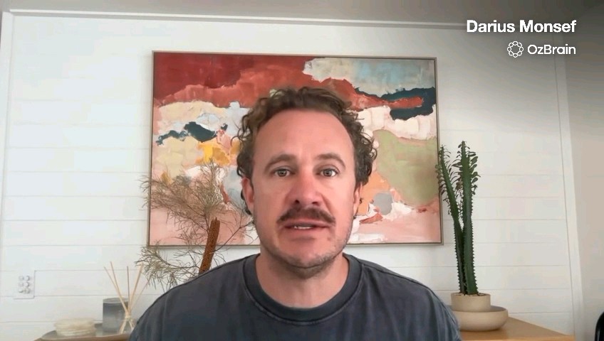 A man with a mustache speaks on a video call labeled 'Darius Monsef' with an 'OzBrain' logo, sitting in front of an abstract painting and potted plants.