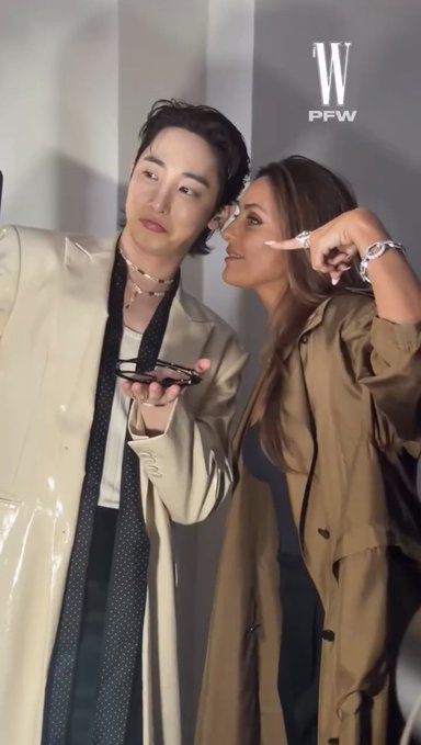 Two women pose closely together at what appears to be Paris Fashion Week, marked by the 'W PFW' watermark; one wears a glossy beige coat with a polka-dot tie, while the other wears a tan trench coat.