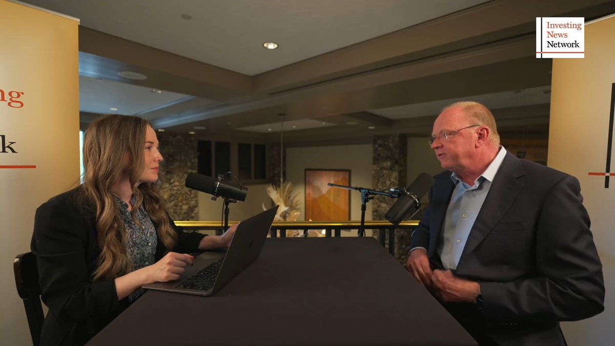 An Investing News Network interview setting with a woman using a laptop interviewing a bald man in a dark suit across a table with microphones.