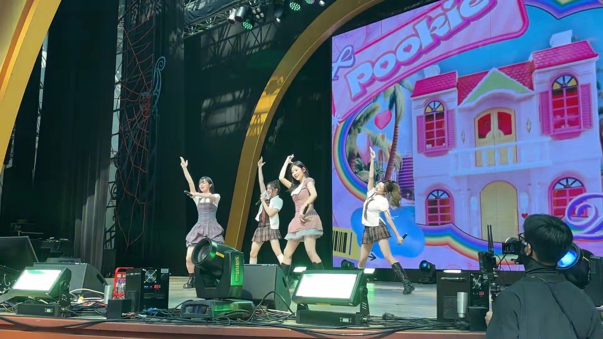 Five young women perform on stage at what appears to be an anime/J-pop convention, with a colorful backdrop displaying 'Poolkie' and a cartoon mansion design.