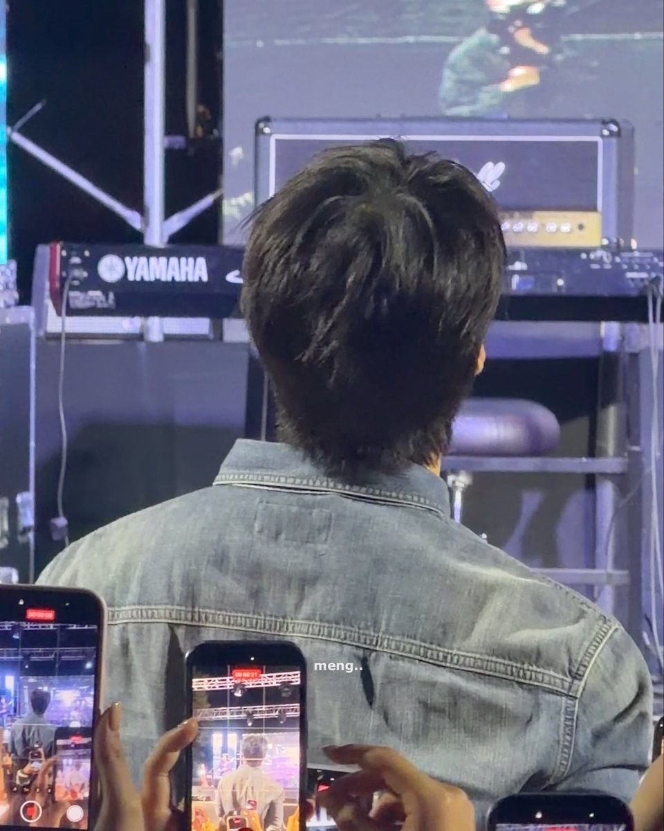 A view from behind a person in a denim jacket facing a Yamaha keyboard on stage, while audience members record with the word 'meng..' overlaid on the image.