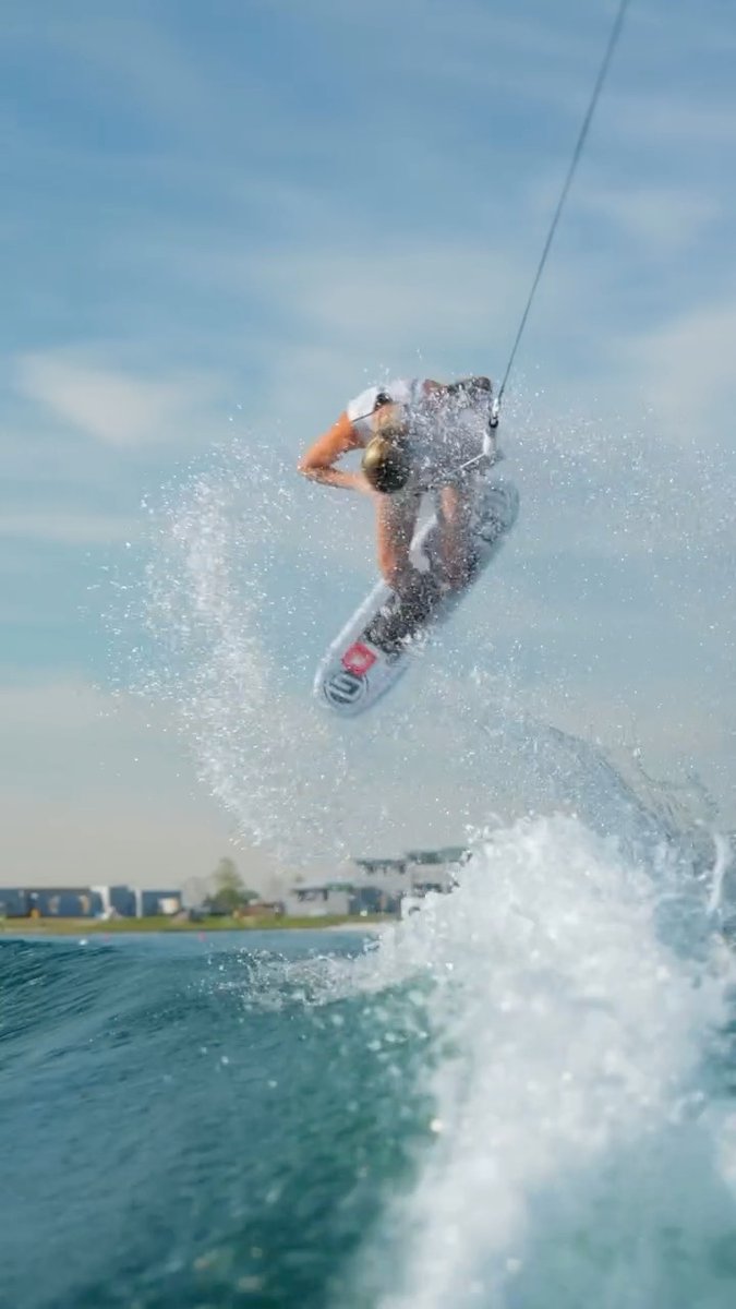 A wakeboarder performing an aerial trick mid-jump above water, splashing against a backdrop of blue sky and distant shoreline.