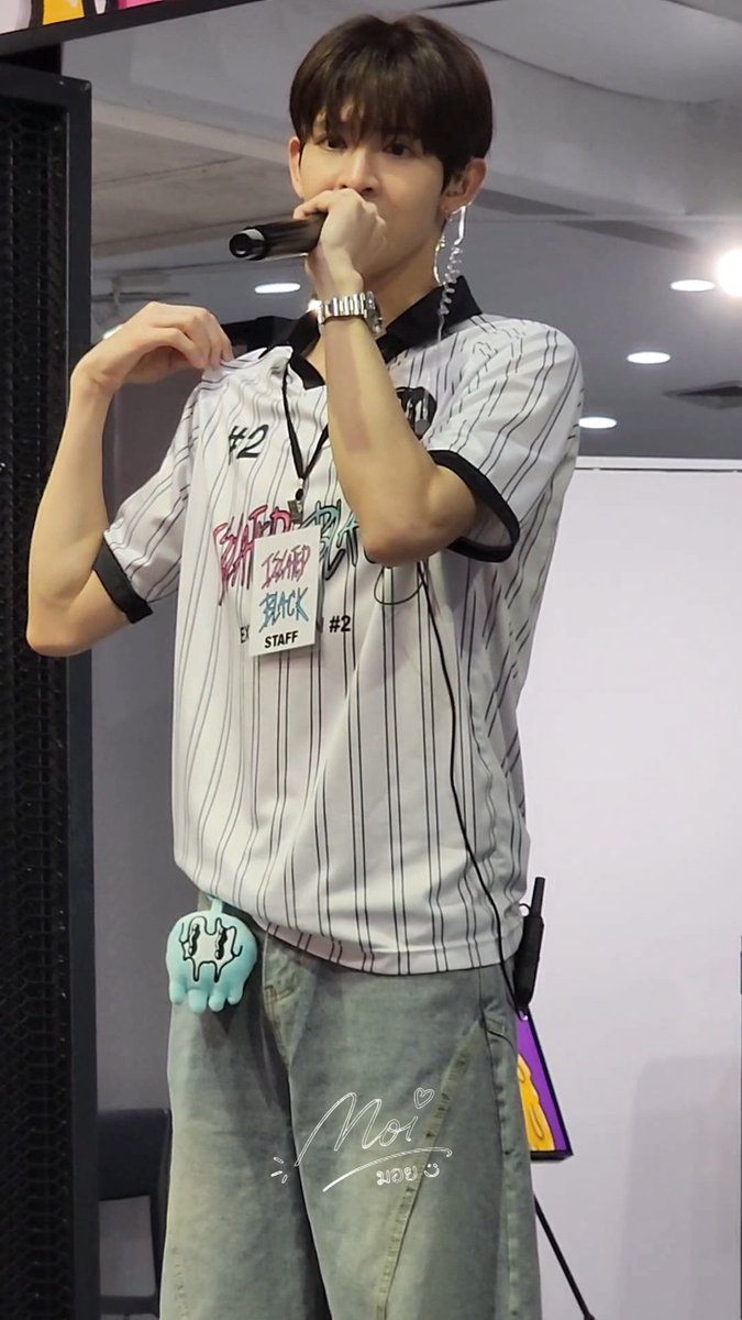 A young man performing with a microphone, wearing a white striped short-sleeve shirt with '#2' graphics, a 'STAFF' lanyard, baggy denim jeans, and a small blue plush toy hanging from his pocket.