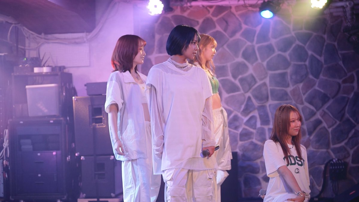 Four young women stand on a dimly lit stage with purple lighting, one holding a microphone, with stone-patterned walls and stacked amplifiers visible in the background.