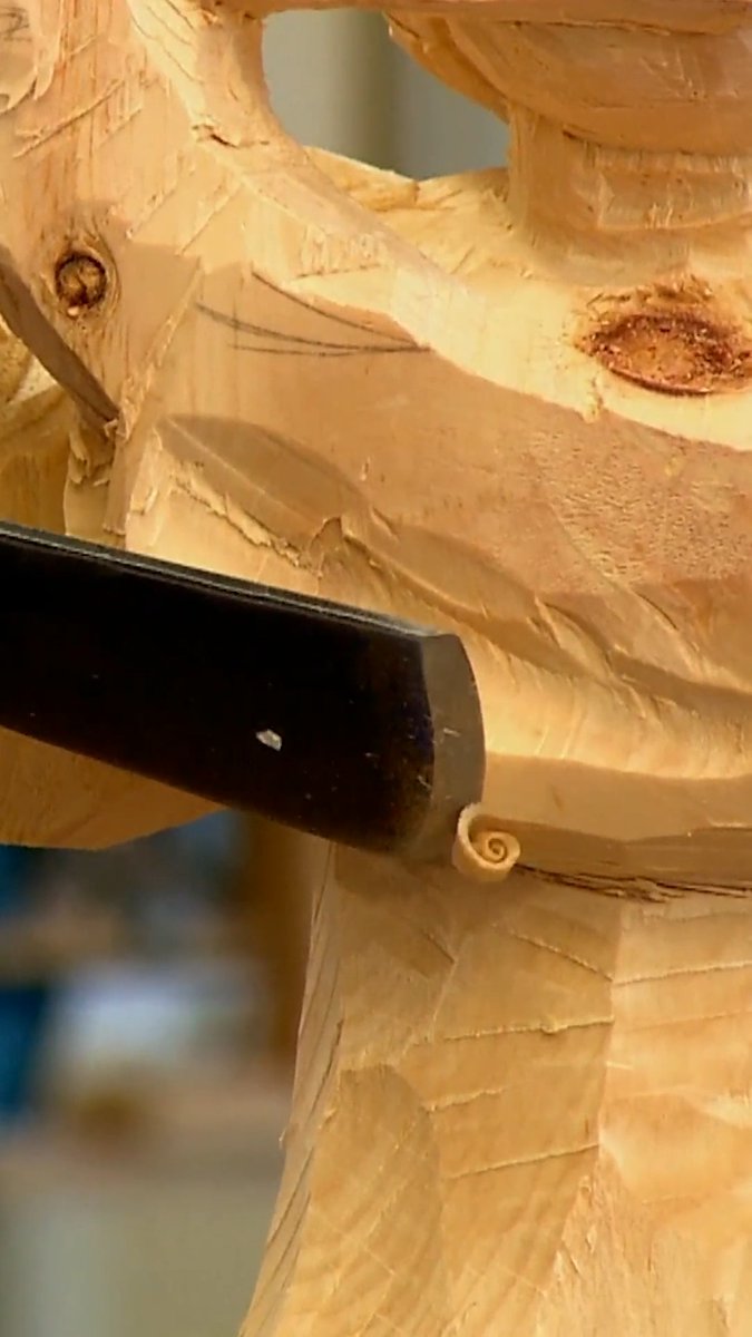 A wood carving chisel shapes a delicate spiral detail into a wooden sculpture, creating fine wood curls as it cuts into the surface. The image perfectly captures the oddly satisfying craftsmanship showcased in How It's Made.