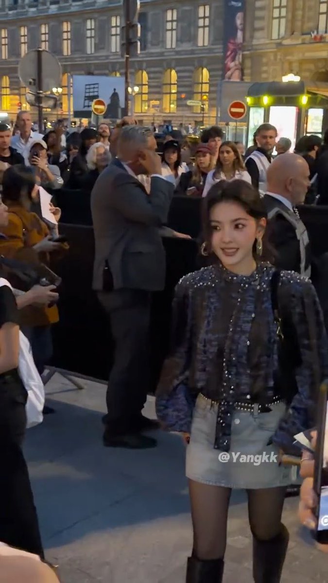 A young woman, possibly a celebrity given the crowd and photographers, poses outdoors at night wearing a blue sequined top, denim shorts, and black tights, with the watermark '@Yangkk' visible.