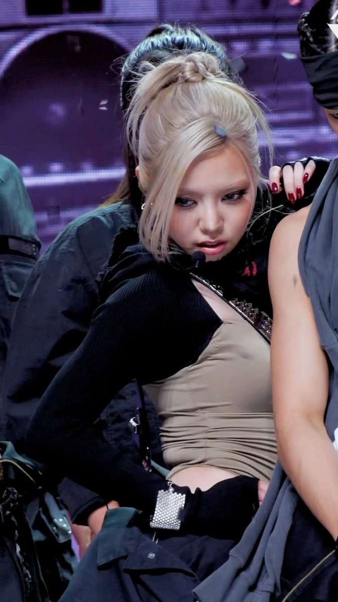 A blonde K-pop performer on stage wearing dramatic eye makeup, a headpiece, and a layered outfit with a black jacket and beige top.