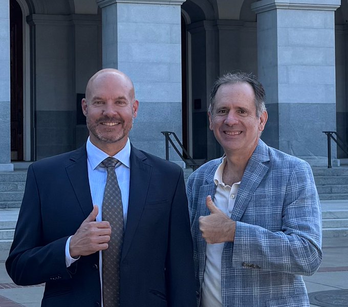 Two smiling men in business attire—one bald with a goatee in a dark suit and tie, the other with gray hair in a blue plaid blazer—both giving a thumbs-up in front of a building with stone columns.