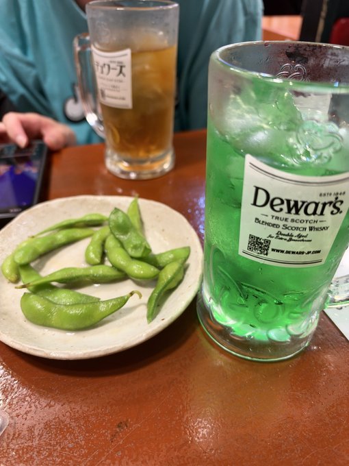 A glass of iced green soda served in a mug branded with the Dewar's True Scotch Whisky logo sits next to a plate of edamame beans and a glass of iced black tea, likely at a Japanese restaurant.