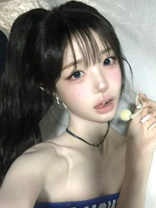 A young woman with long dark hair in a ponytail poses for a selfie, holding a lollipop, wearing a blue strapless top and a thin black choker.
