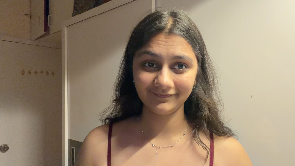 A selfie-style photo of a young woman with long dark hair, wearing a maroon top and a delicate gold necklace, smiling softly in a room with white cabinets.