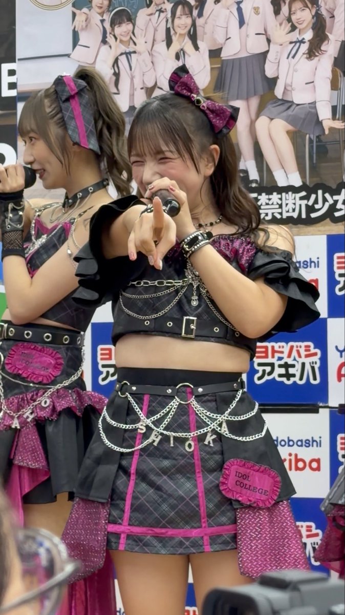 Two young women in matching black plaid and pink idol outfits with 'IDOL COLLEGE' patches perform on stage, with the front performer pointing at the camera; the backdrop features a photo of the group and Japanese text including 'ヨドバシ アキバ'.