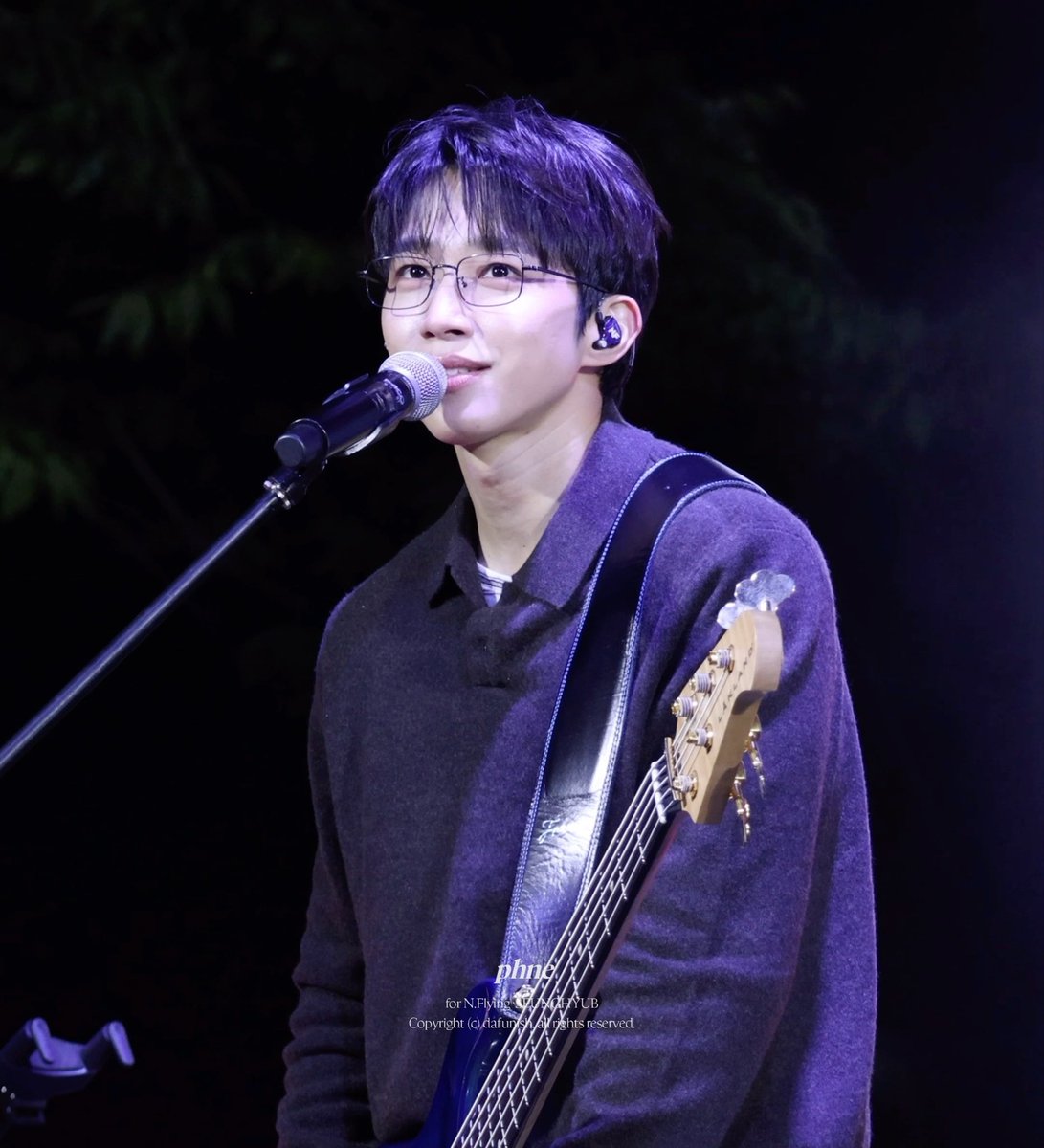 An Asian male musician wearing glasses, a dark sweater, and an in-ear monitor stands at a microphone while holding a blue bass guitar on a dimly lit stage.