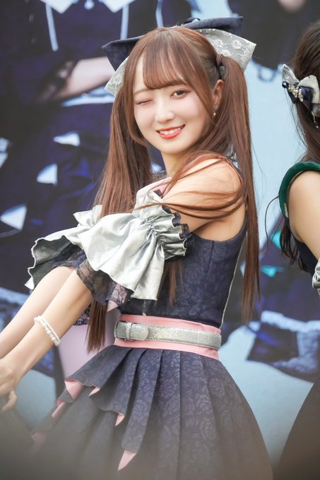 A smiling young woman with long brown hair in pigtails winks at the camera while wearing a black and pink dress with a bow.
