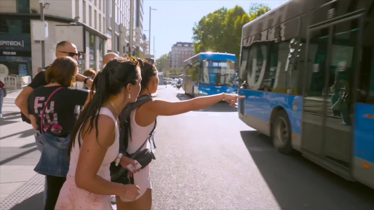 A group of pedestrians stands on a city street corner as a woman points toward a passing blue city bus with route text "316.SAN NICOLA".