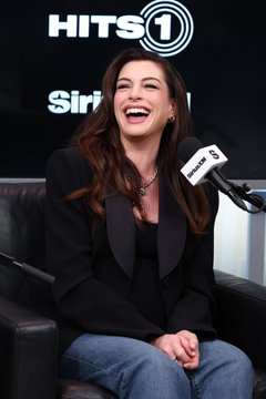 Actress Anne Hathaway laughs joyfully while sitting in a chair during a SiriusXM Hits 1 radio interview, with a microphone visible in the foreground.