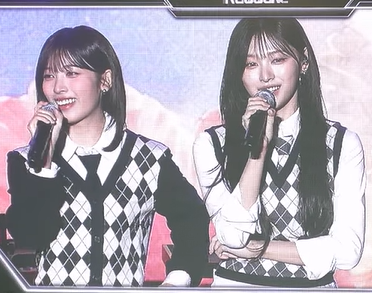 Two young women with long dark hair, wearing matching black and white argyle vests over white shirts, smile while holding microphones during what appears to be a live performance.
