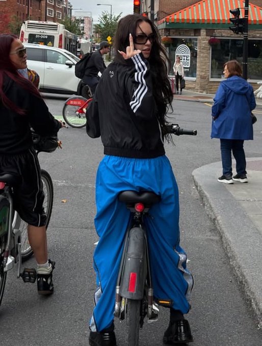 A person with long dark hair and sunglasses rides a bike on a city street, wearing a black Adidas jacket and blue track pants while flashing a peace sign.