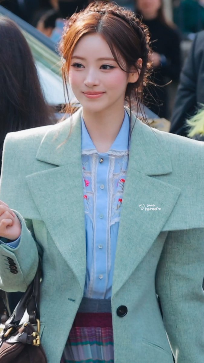 A young woman with her hair tied back smiles softly, wearing a light blue collared shirt under an oversized mint green blazer; a small watermark reads 'juno_tired♡'.