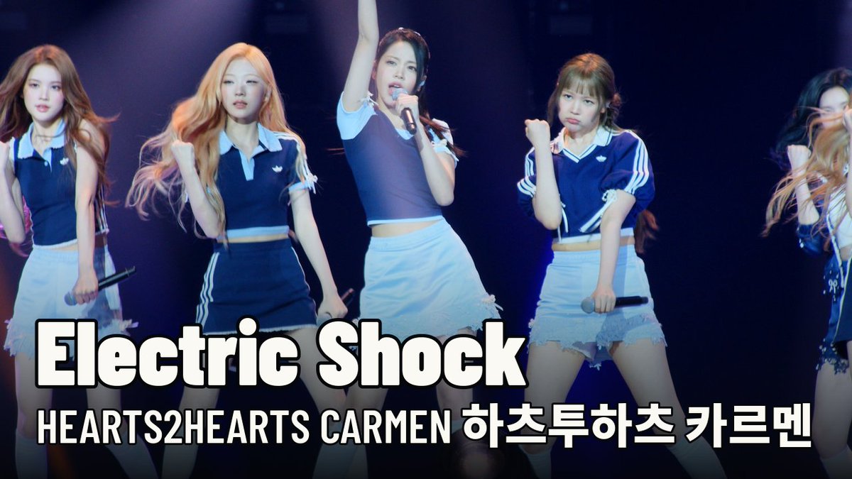Members of the K-pop group HEARTS2HEARTS perform 'Electric Shock' on stage, with center member Carmen highlighted in white, and text reading 'HEARTS2HEARTS CARMEN 하츠투하츠 카르멘' displayed below.
