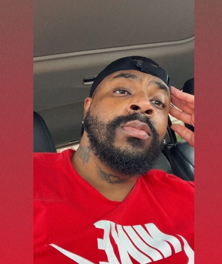A man with a beard wearing a backwards black cap and a red shirt takes a selfie inside a car.
