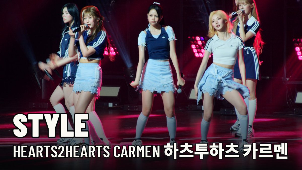 Five female K-pop performers dance on stage in coordinated schoolgirl-inspired outfits with blue vests and white skirts; text overlay reads 'STYLE HEARTS2HEARTS CARMEN 하츠투하츠 카르멘'.