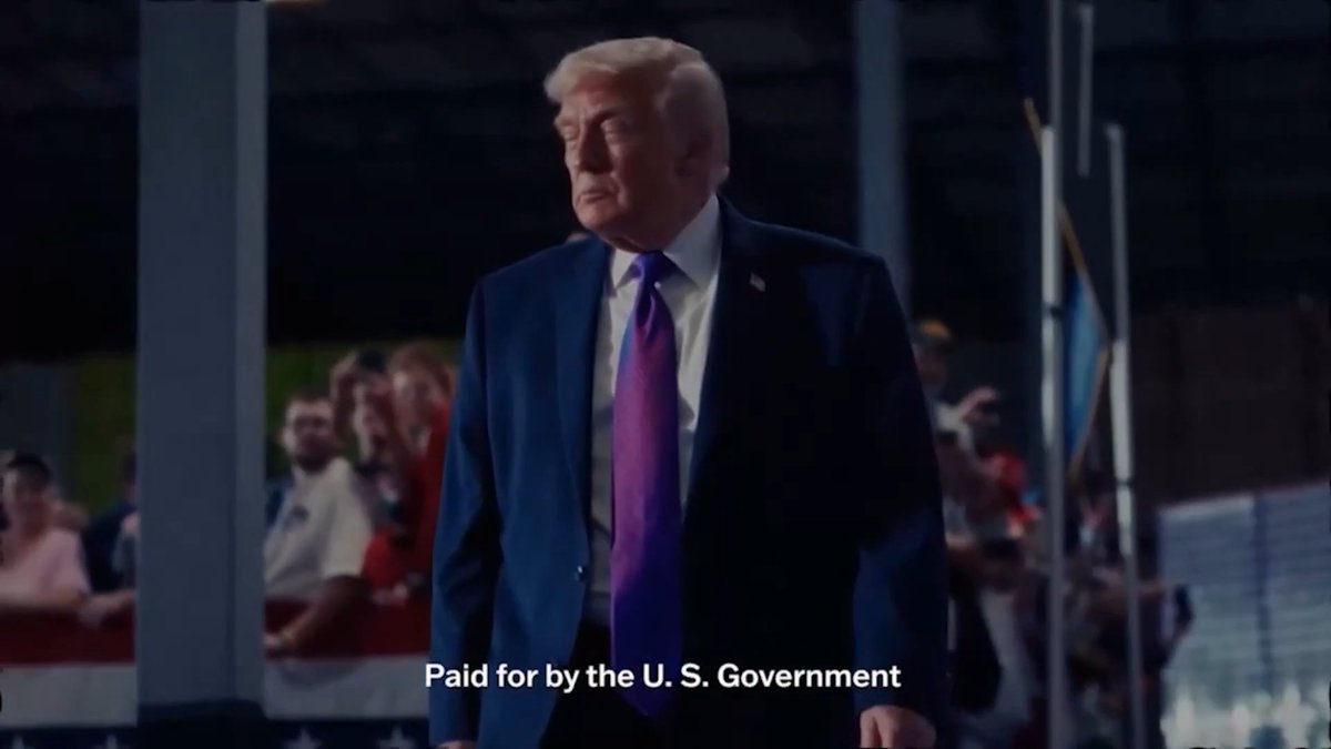 A man with blonde hair wearing a dark suit and purple tie stands at what appears to be a rally, with a crowd visible in the background. The text at the bottom reads "Paid for by the U. S. Government," illustrating the context of the accompanying post about $3.5 million in taxpayer dollars being spent on pro-Trump midterm ads.