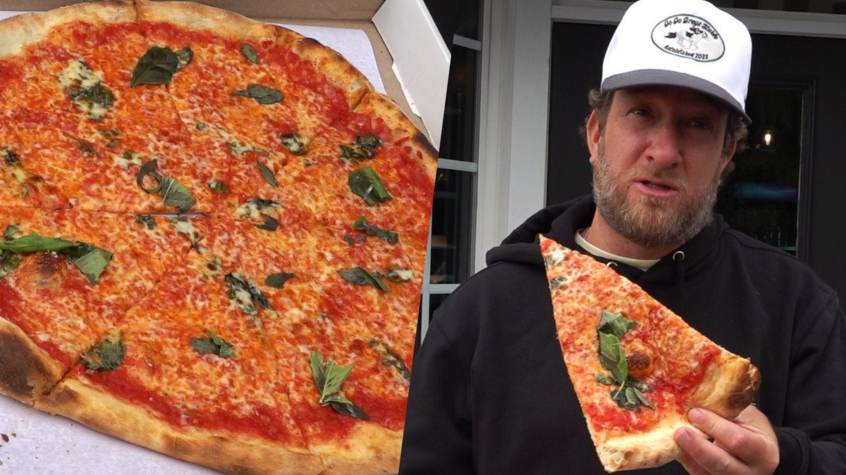 A large Margherita pizza topped with red sauce, melted cheese, and fresh basil sits in an open box beside a bearded man wearing a black hoodie and white cap reading "Da Da Great Black," who holds up a triangular slice for the camera as part of a Barstool Pizza Review of Mommò Pizza Shop in Fishkill, NY.