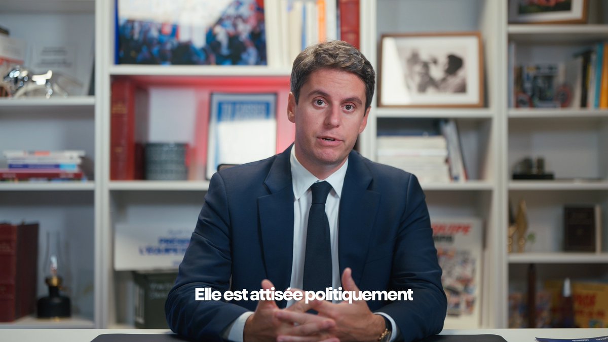 A man in a navy suit sits at a desk in front of a bookshelf, speaking directly to the camera with the French subtitle "Elle est attisée politiquement" ("It is being stoked politically") displayed at the bottom of the frame, reinforcing his message about the need to restore calm and dialogue amid politically charged tensions.