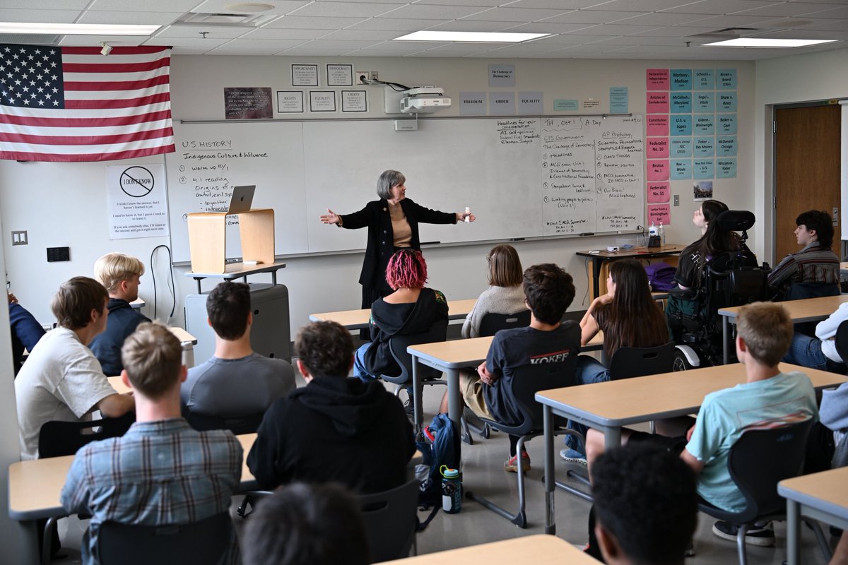 A teacher stands at the front of a classroom, gesturing toward seated students. A large whiteboard with handwritten notes and a U.S. flag are behind her. There is text on the board and classroom displays.