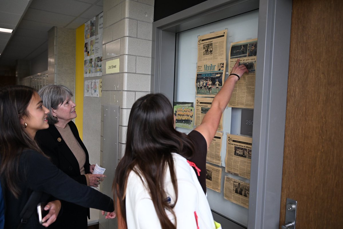Three people stand in a school hallway looking at a display case filled with newspaper clippings. One person points to an article. There is text on the clippings and signs.