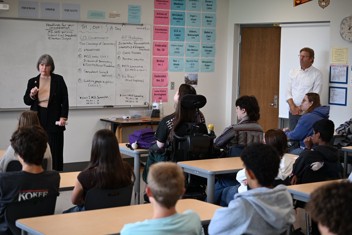 A classroom scene with a woman teaching at the front while students sit at desks and listen. A man stands near the door. The whiteboard and walls have schedules, notes, and educational materials.