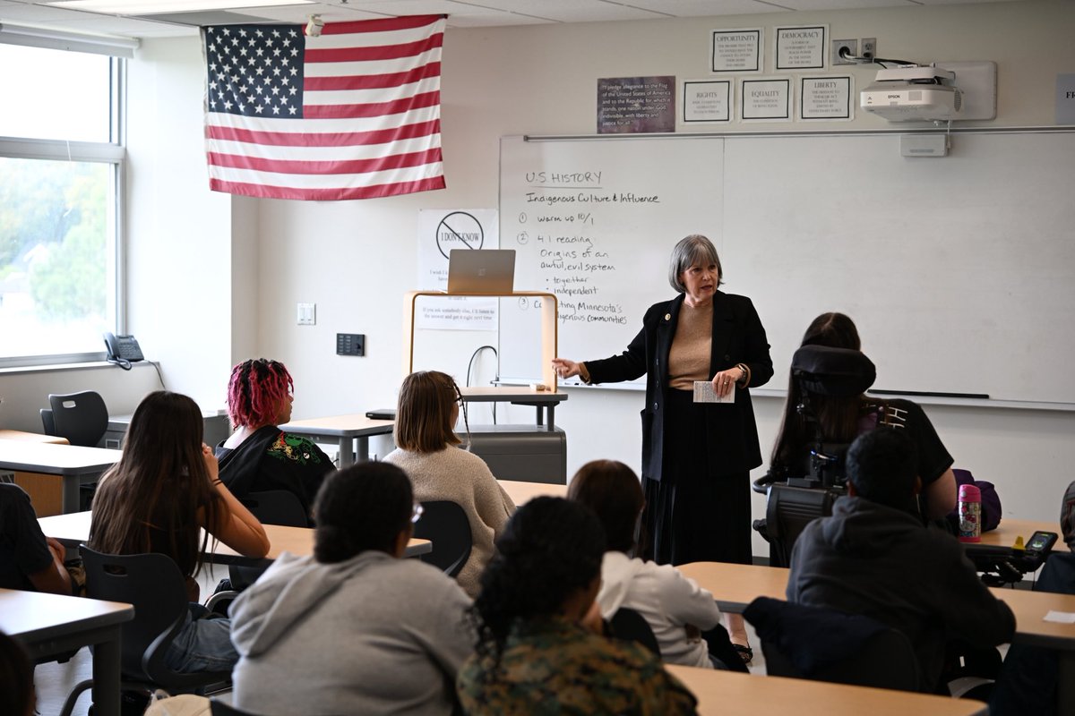 A teacher speaks to students seated at desks in a classroom. An American flag hangs near the windows, and a whiteboard with handwritten notes and classroom equipment is visible behind the teacher. There is text on the board and wall displays.