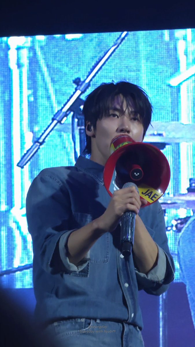 A performer holding a red megaphone with "JAE" written on it speaks into a microphone during a concert, with blue stage lighting in the background.