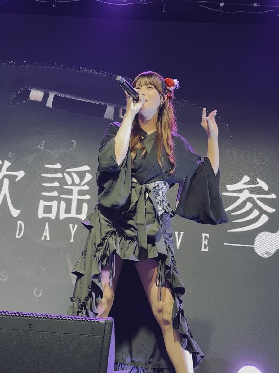 A photograph of a woman performing on stage, wearing a black dress with ruffled skirt, holding a microphone and singing, with Chinese characters visible on the screen behind her.
