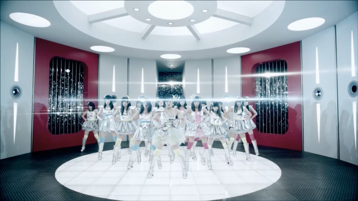 A group of performers in shiny silver costumes strike a pose on a circular platform in a futuristic studio with red and white decor. The image visually represents the music video or promotional content for NMB48's 8th single "Kamonegix," which celebrated its 13th anniversary as noted in the caption.