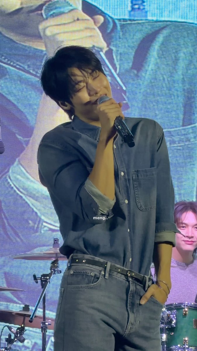 A man in a dark denim shirt sings passionately into a microphone on stage, eyes closed, with drum equipment visible and another person in the background; a small caption 'meng..' is overlaid.